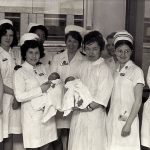 Photograph of 8 nurses in white uniforms and hats posing for the camera. The two midwives in the centre are each holding a new-born baby