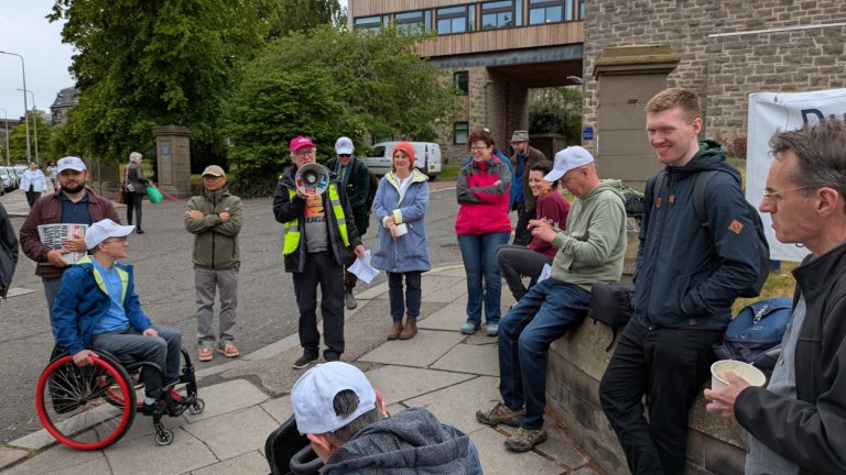 A group of people stand and sit on a wall outside the University of Dundee Tower Building. They are listening to a man speak through a loud hailer.